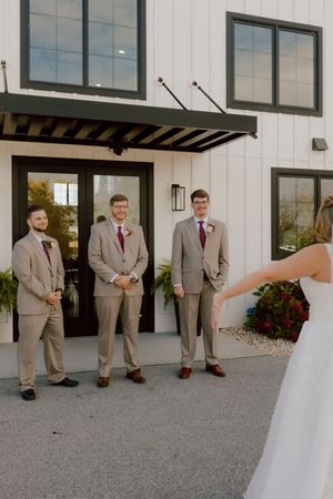Bride approaches three groomsmen in tan suits with burgundy ties outside a white venue.