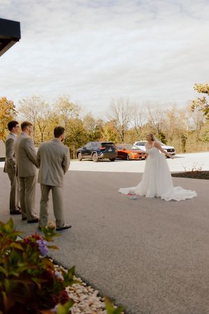 Bride in white gown approaches groomsmen in tan suits outside a wedding venue with cars in background.