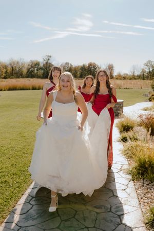 Bride in white ball gown walks with three bridesmaids in red dresses outdoors.