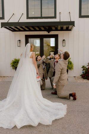 Groomsmen kneel and celebrate with drinks as bride in white gown and veil stands before white venue.