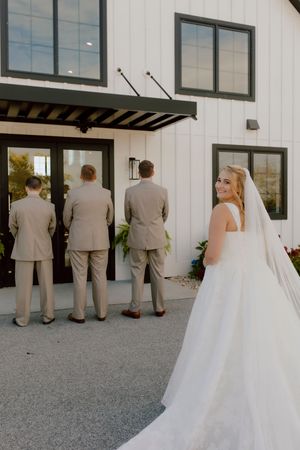 Bride in white gown smiles as groomsmen in tan suits face a white barn venue.