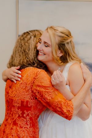 Smiling bride in white dress sharing warm hug with woman in orange lace dress.