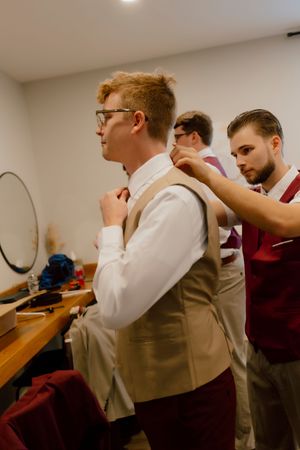 Groomsmen help each other get dressed in tan vests and white shirts inside a wedding preparation room.