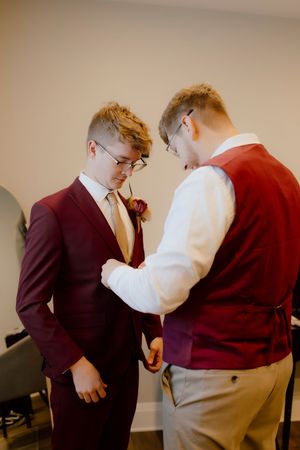 Groom in burgundy suit adjusts boutonniere with help from groomsman in tan vest before wedding ceremony.