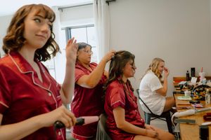 Bridesmaids in red robes get hair styled at mirrors during wedding morning preparations indoors.