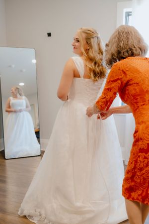 Bride in white gown getting dress fastened by woman in orange lace dress near mirror.