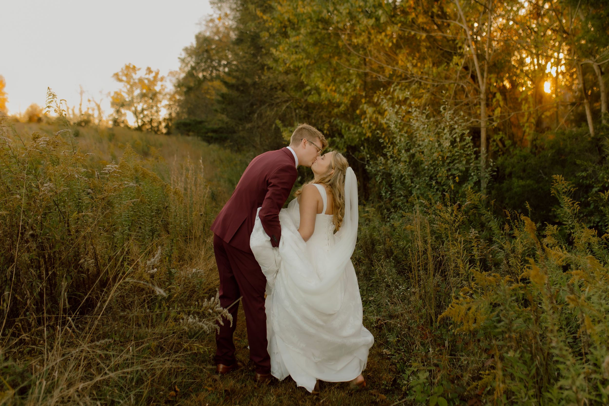 Bride and groom share a kiss at golden hour in a lush green field.
