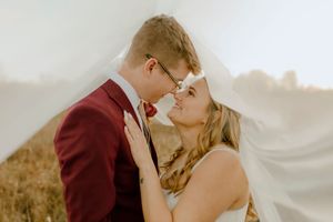 Romantic bride and groom almost kissing under flowing veil in sunlit field.