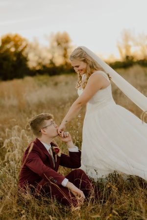 Groom sits in golden meadow kissing bride's hand while she smiles down at him.