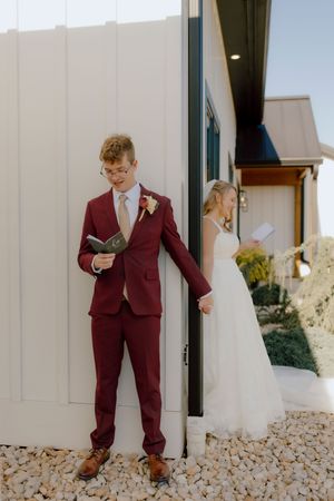 Groom in burgundy suit and bride in white gown reading private vows before the ceremony.