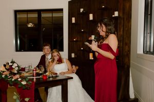 Bridesmaid in red dress giving speech at wedding reception while couple sits at table.