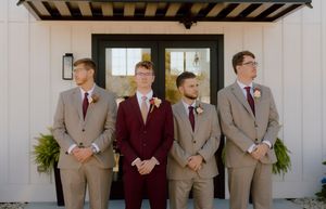 Four groomsmen in tan and burgundy suits with boutonnieres pose outside a modern white wedding venue.