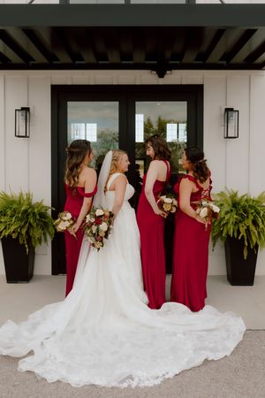 Bride in white gown and veil stands with three bridesmaids in red dresses holding bouquets outdoors.