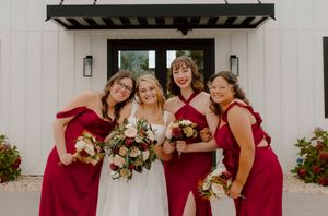Smiling bride with bouquet poses with three bridesmaids in red dresses outside a white wedding venue.