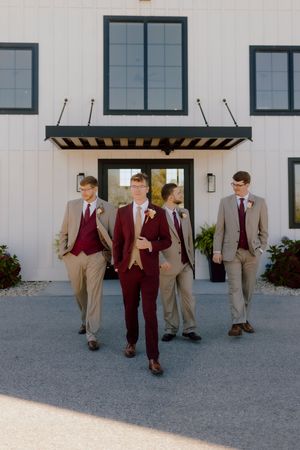 Groom in burgundy suit leads three groomsmen in tan suits walking confidently outside white venue.