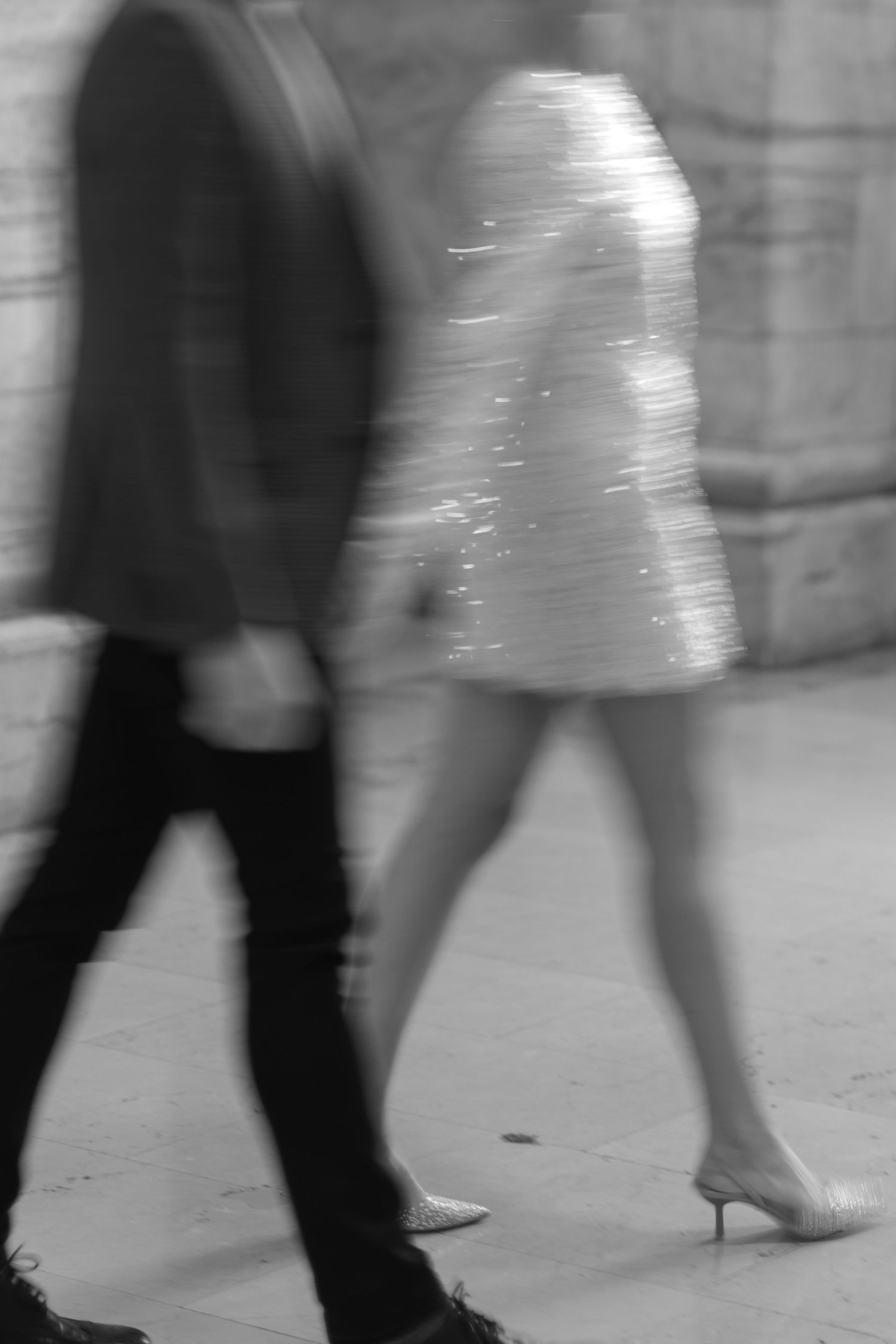 Artistic motion-blurred black and white photo of a couple walking past a stone wall, woman in a short dress.