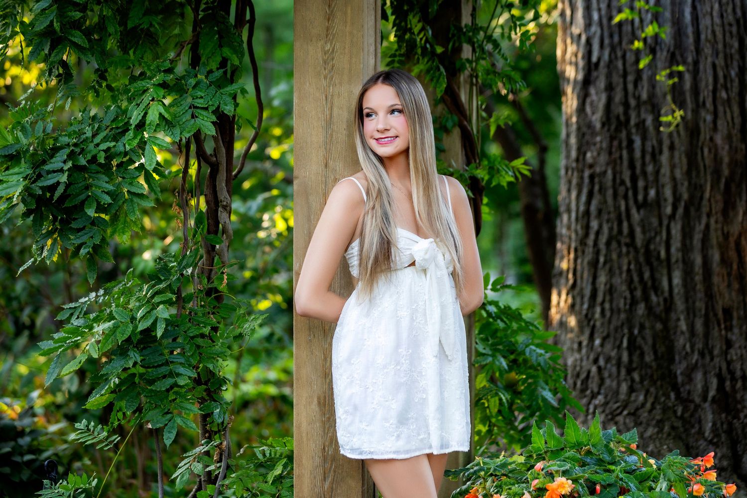 Person in white jacket and dress poses between garden archways with green foliage.
