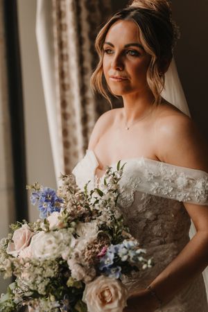 A bride in an off-shoulder lace gown holds a romantic pastel bouquet with roses and delphinium flowers.
