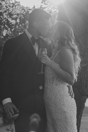 Black and white image of couple sharing intimate moment with champagne glasses at outdoor wedding celebration in sunlight.
