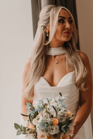 Bride in white dress poses with delicate blue and white wedding bouquet in soft indoor lighting.