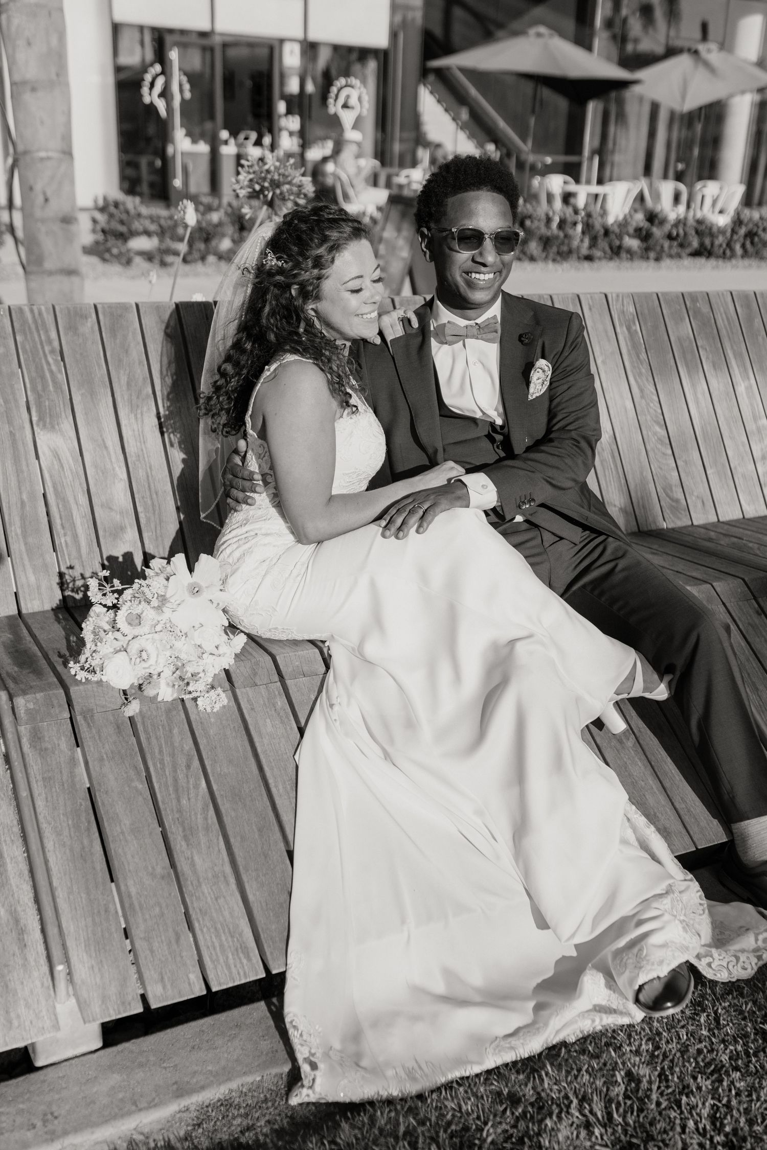 A man and woman in wedding attire looking away and smiling sitting on a bench.