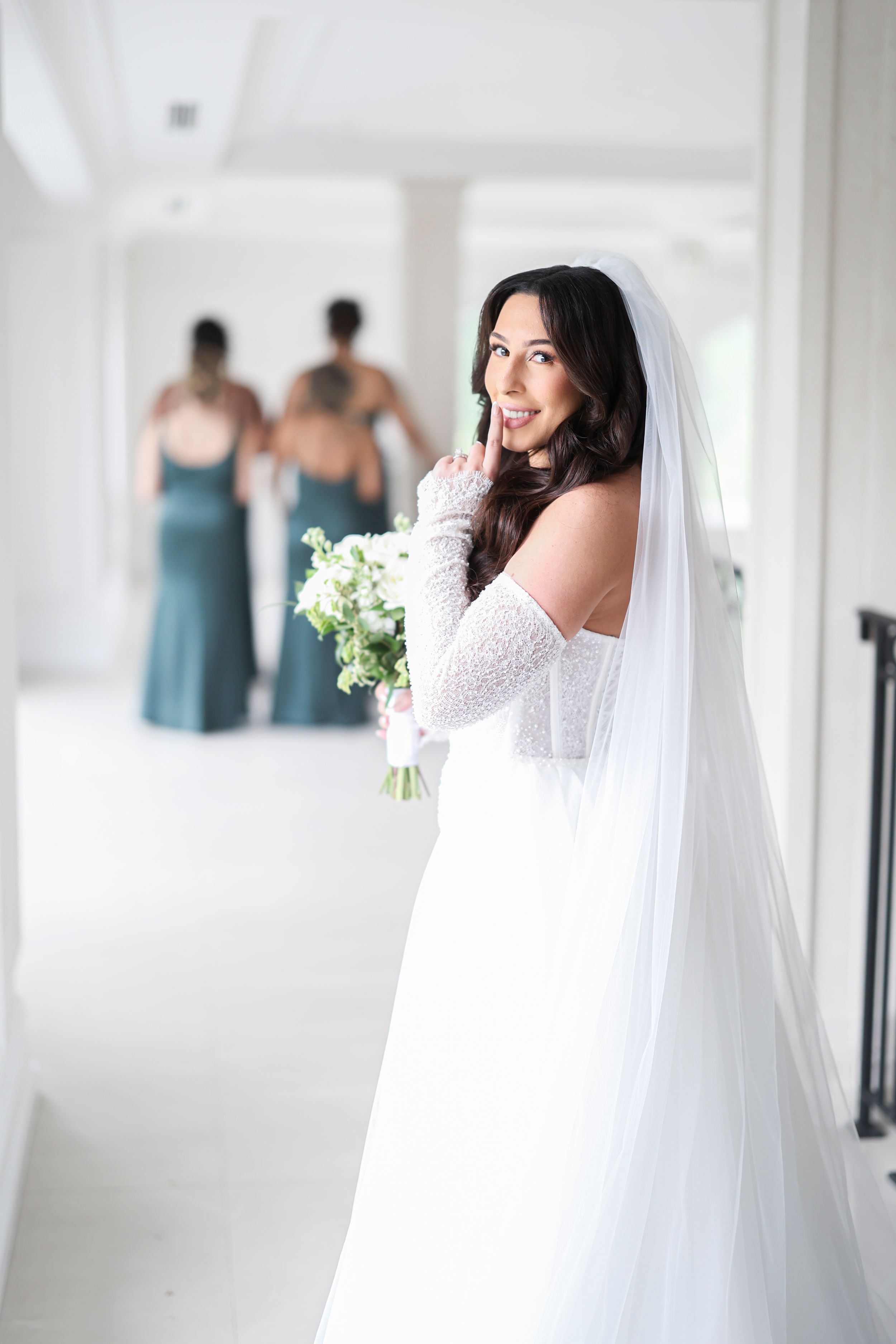A bride in a white wedding dress and long veil poses elegantly in a bright, airy room with turquoise bridesmaid dresses visible.