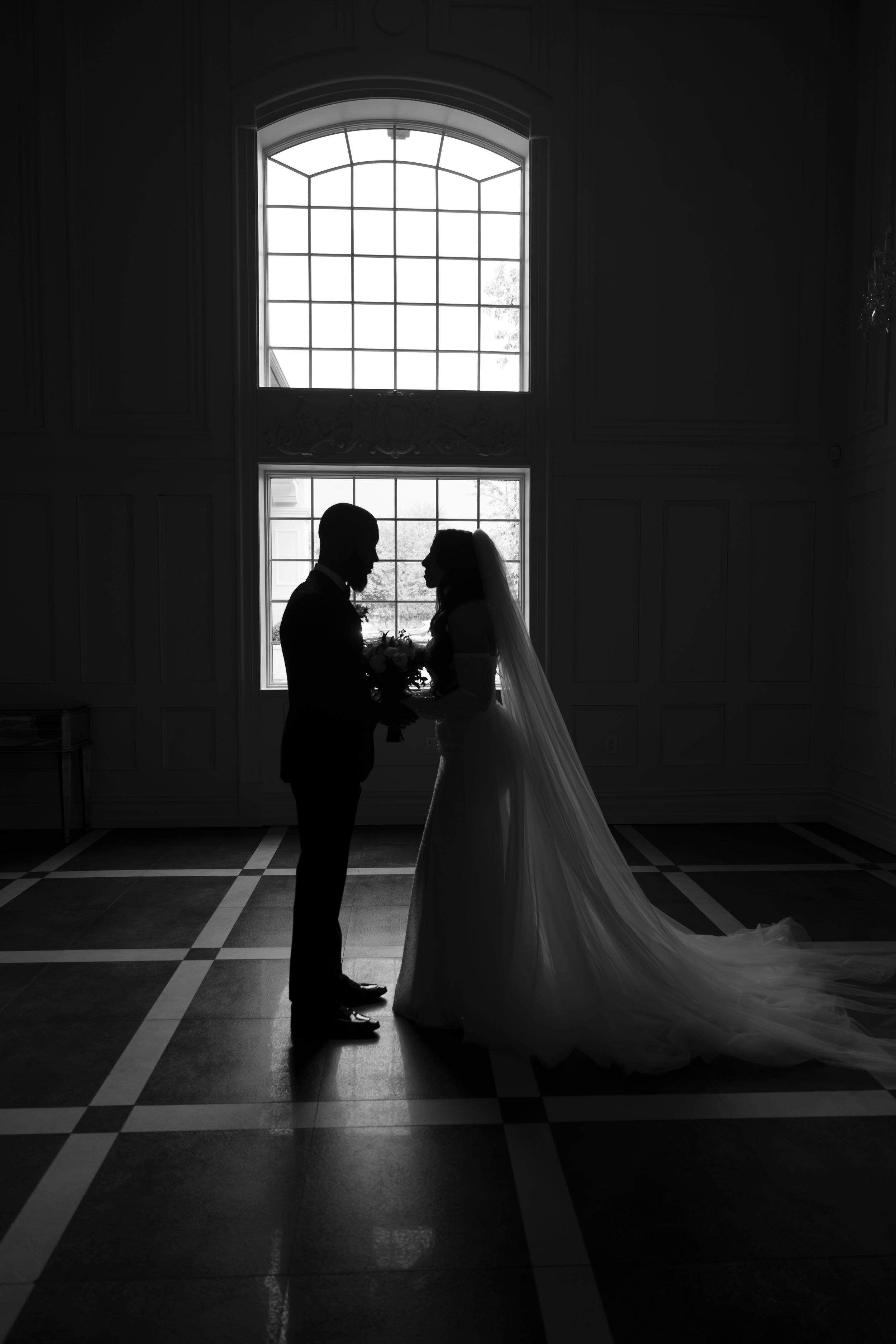 A silhouetted couple stands in front of a large arched window creating a dramatic backlit scene in black and white.