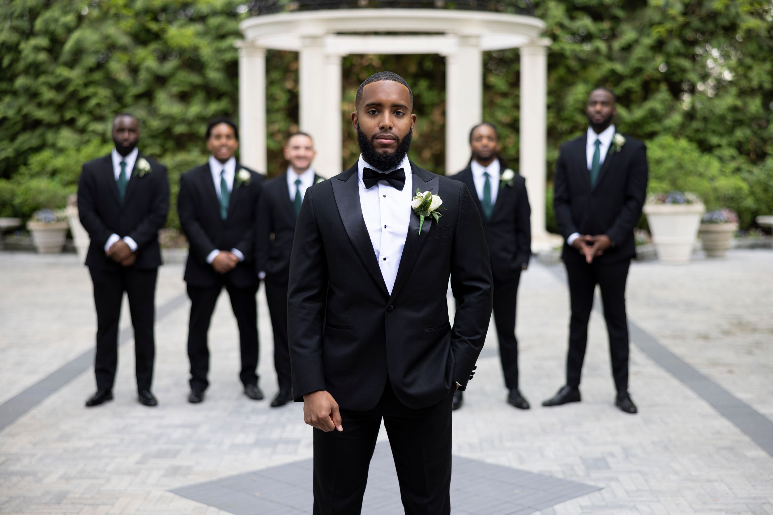 A wedding party in formal black tuxedos poses outdoors in front of a white gazebo structure.
