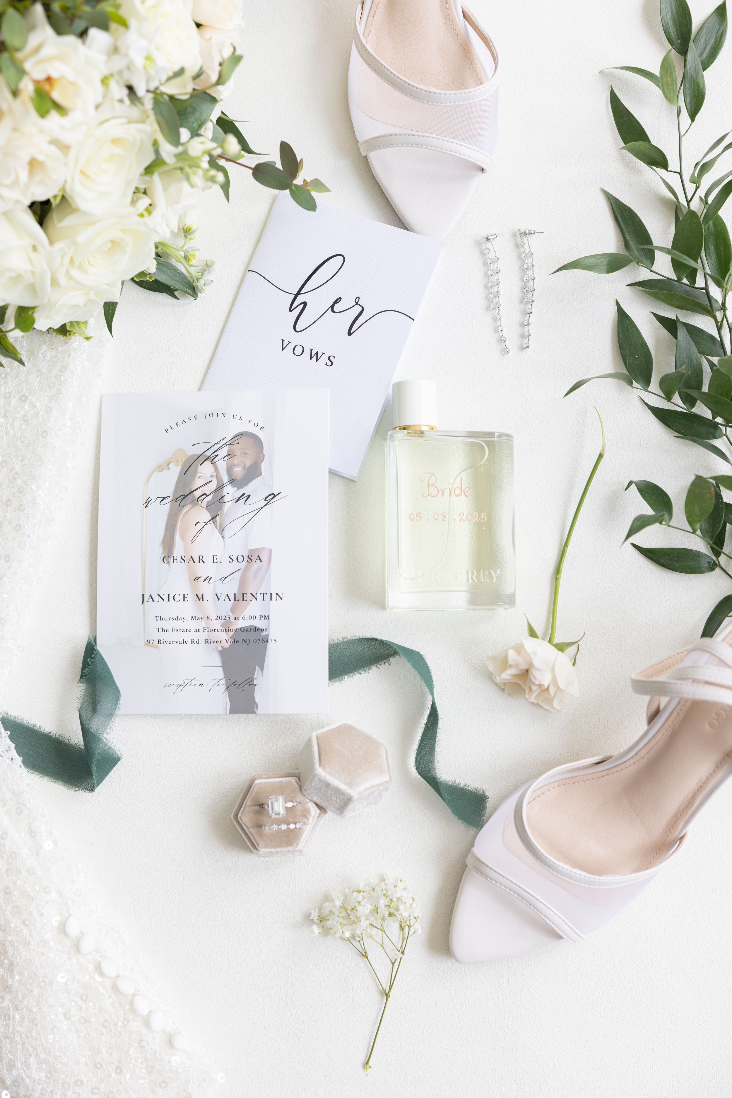 Elegant wedding details flatlay with vow book, white shoes, floral elements and perfume bottle on white background.
