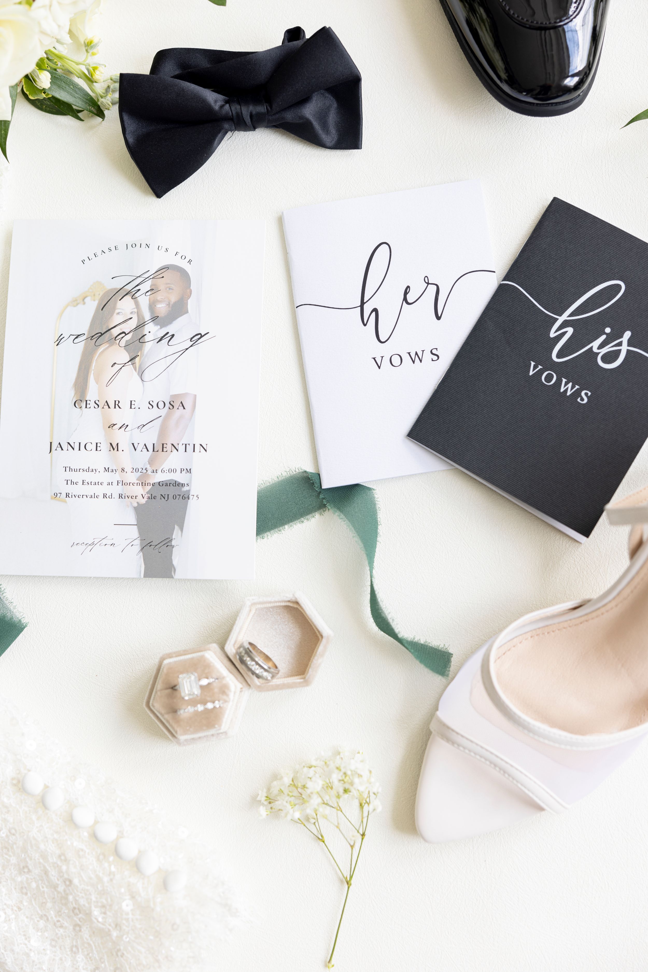Flat lay of wedding details including black bowtie, vow books, rings, invitation and flowers on white background.
