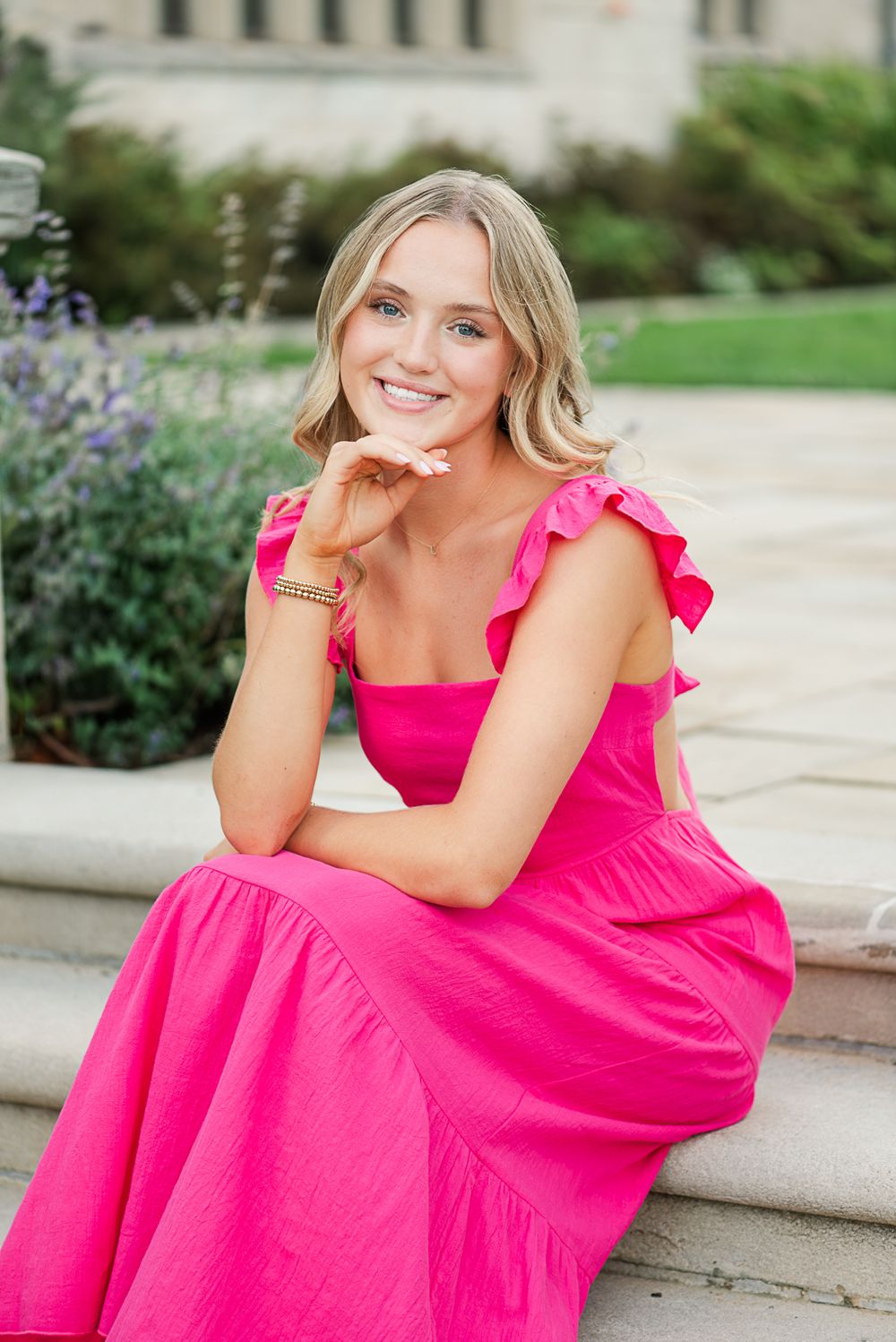 A bright pink maxi dress with ruffled sleeves photographed on outdoor stone steps with purple flowers in the background.