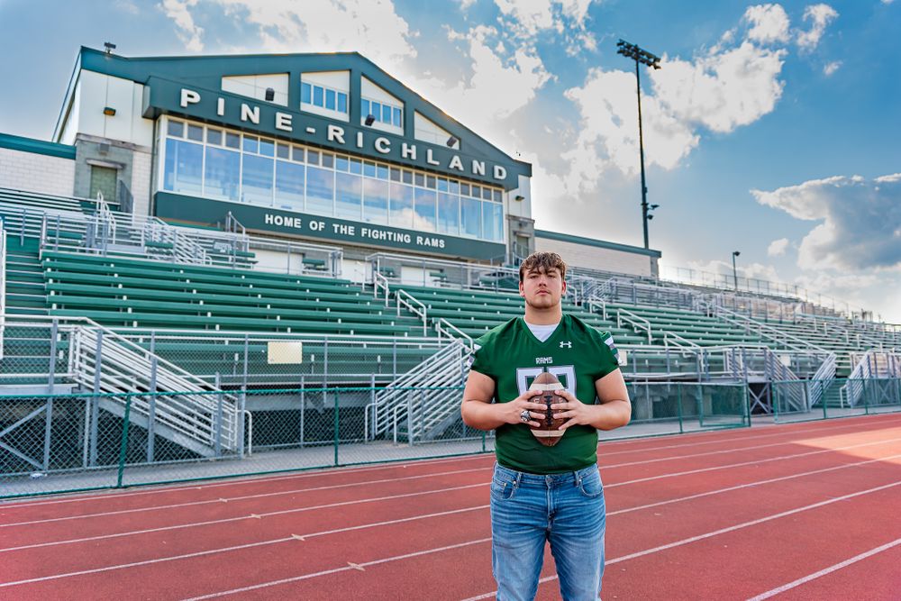 Football player stands on track field in front of green stadium bleachers and press box on sunny day.