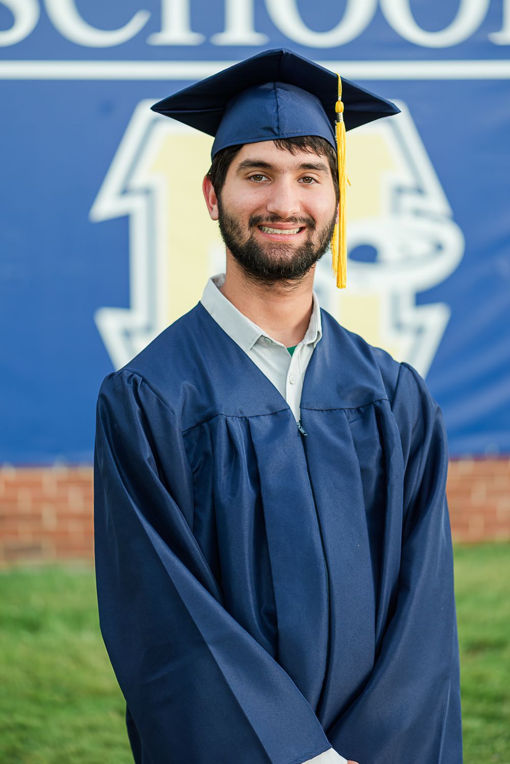 Graduate student in blue cap and gown standing in front of a school logo backdrop.