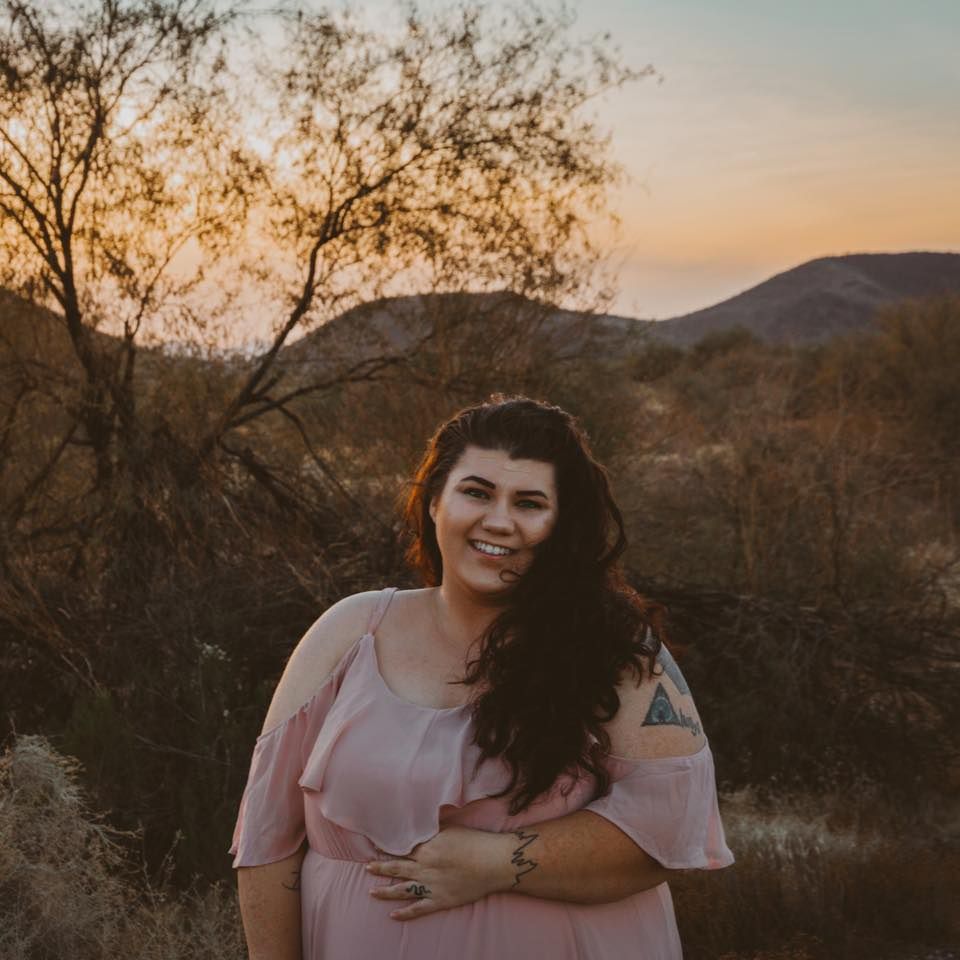 A plus size woman with long brown hair in a pink dress poses outdoors at sunset with mountains and desert brush in the background.