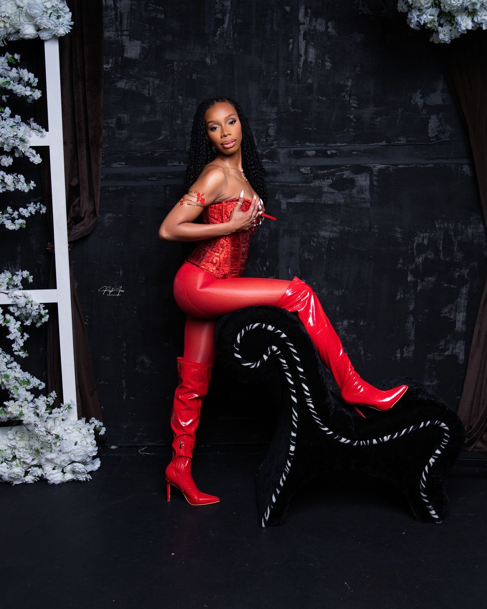 Woman in red corset, shiny red latex pants, and thigh-high red boots posing in a dark studio with white flowers.