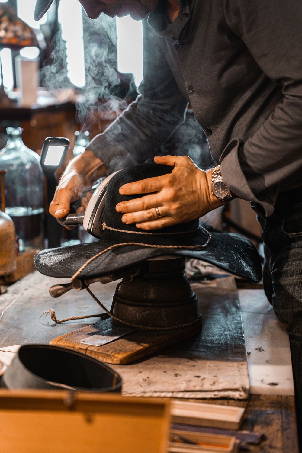 Craftsman steaming and shaping a hat on a vintage hat block in a traditional hat-making workshop.