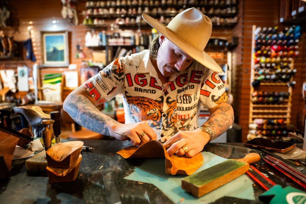 Tattooed craftsman in cowboy hat and tiger shirt works on leather crafting project at a workshop bench.