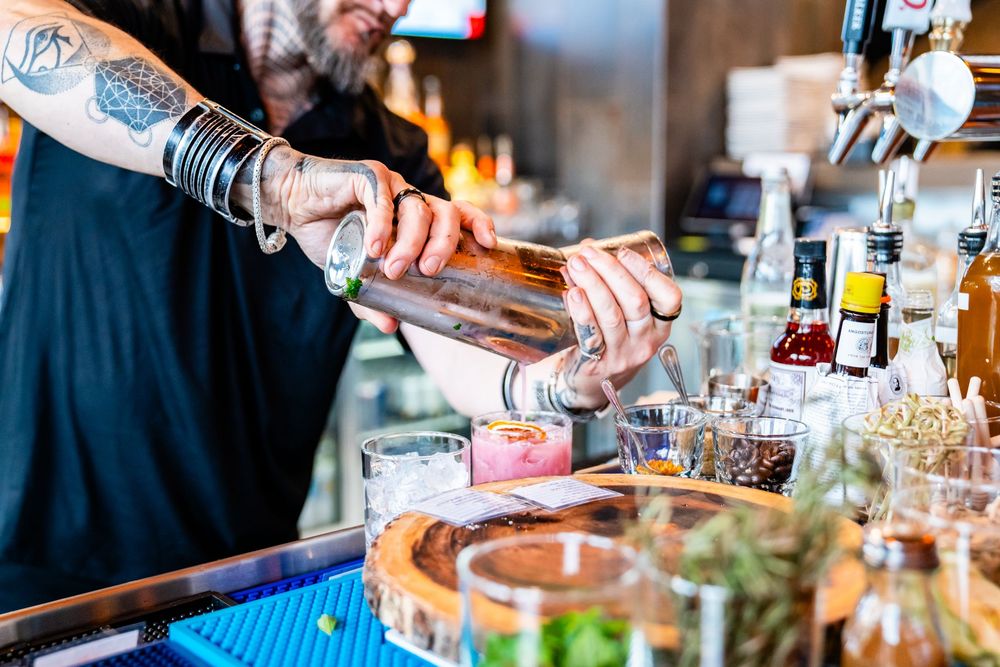 Tattooed bartender pouring a cocktail shaker at a bar with bottles and glasses in the background.