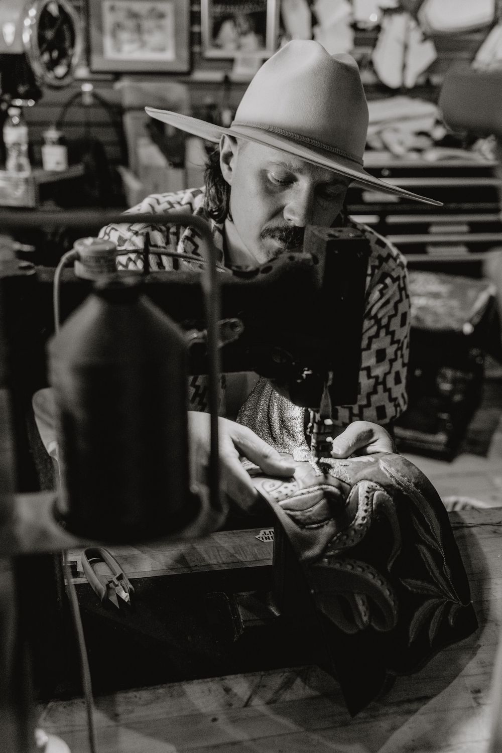 Black and white photo of a craftsman wearing a wide-brim hat using a sewing machine in a workshop.