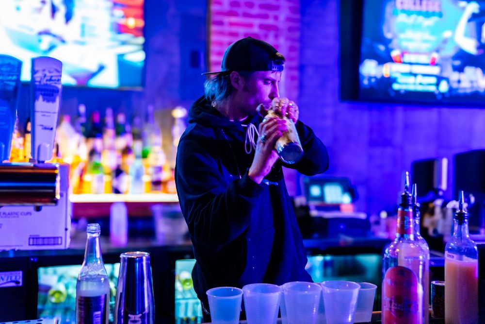 Bartender shaking a cocktail drink behind a neon-lit bar with bottles and cups on the counter.
