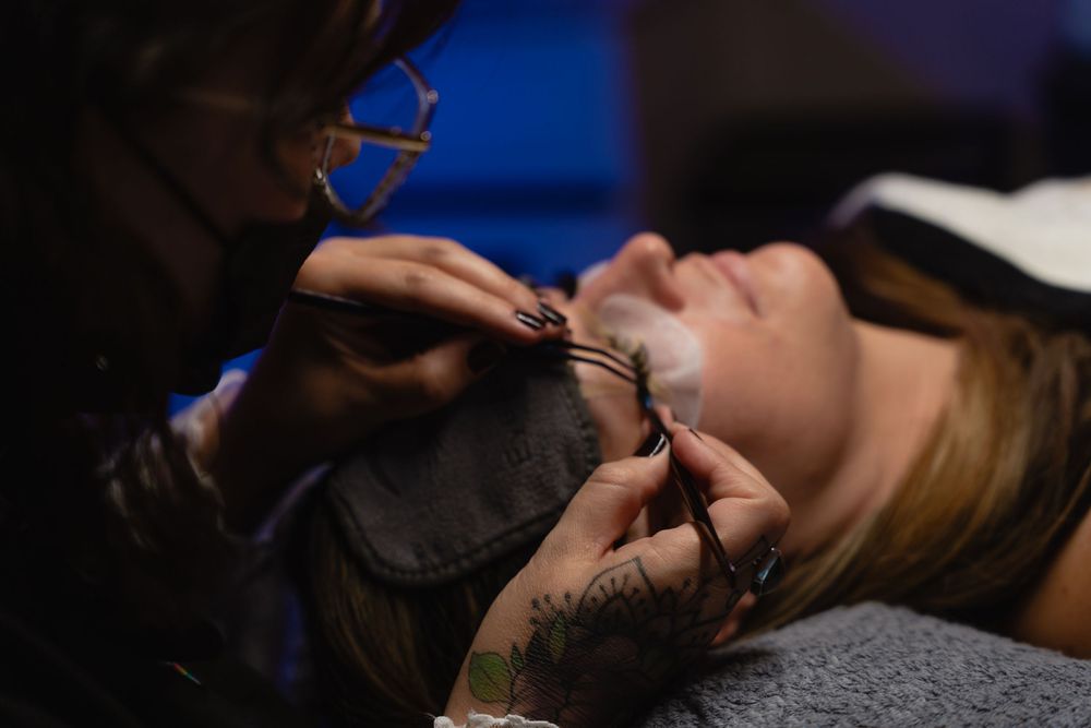 Tattoo artist applying eyelash extensions to a woman lying down in a professional beauty salon.