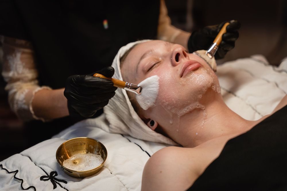 Esthetician applying white facial mask with brush on woman lying on spa treatment table with gold bowl nearby.