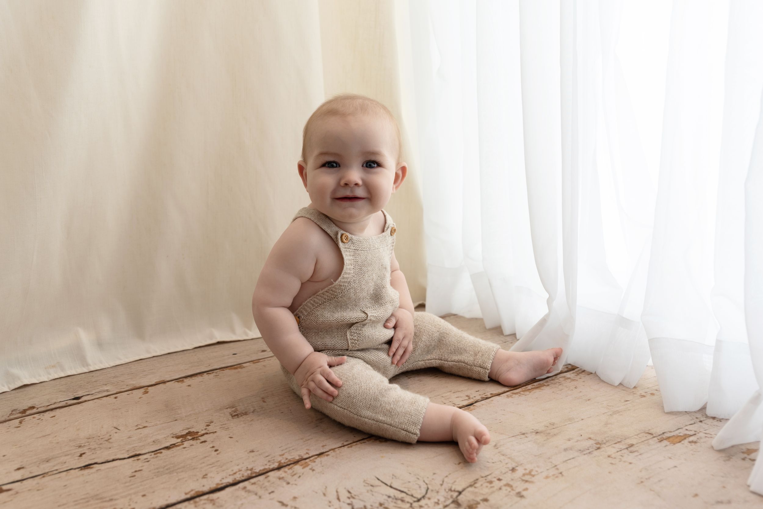 A baby wearing beige linen clothes sits on wooden flooring against white curtains in a minimalist setting.