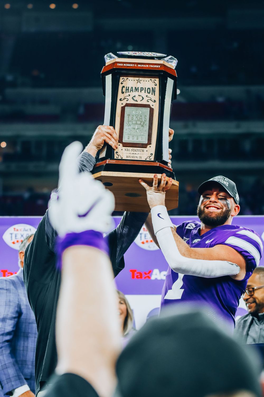 Championship trophy being held up during celebration at football game with purple and white team colors.
