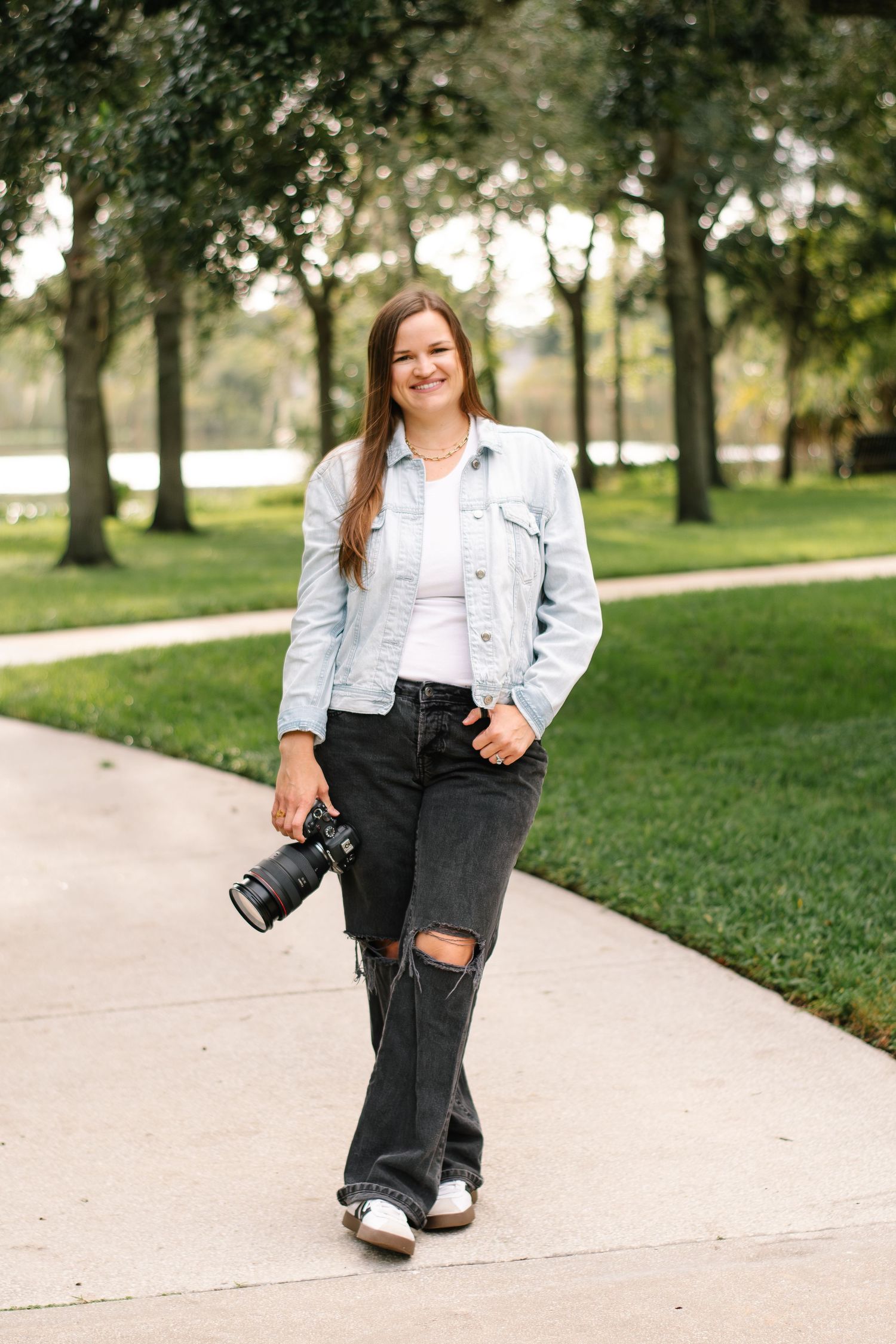 Professional photographer in denim jacket and ripped jeans stands on park pathway with camera.