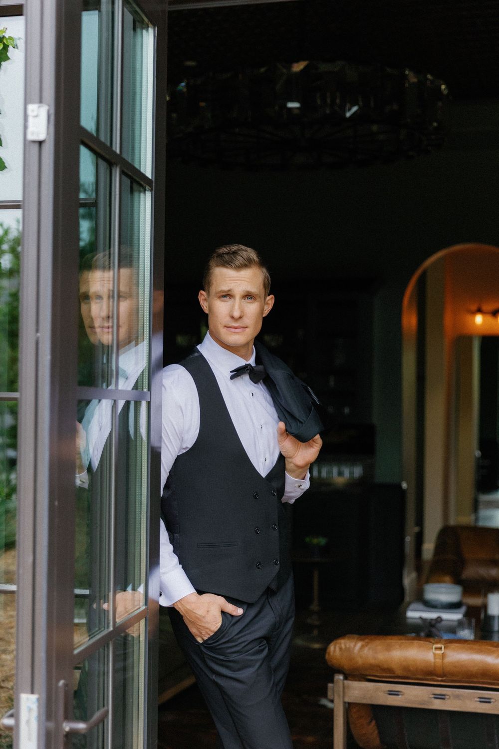Well-dressed server in formal vest and bowtie stands confidently by glass doorway of upscale restaurant.