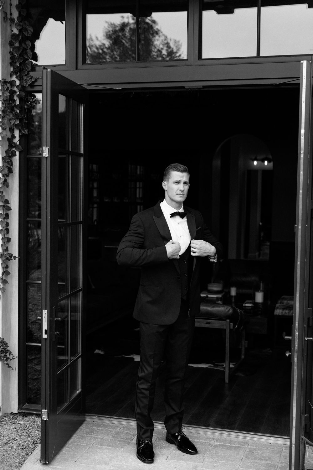 A groom in a classic black tuxedo and bow tie stands in an elegant doorway adjusting his cuff.