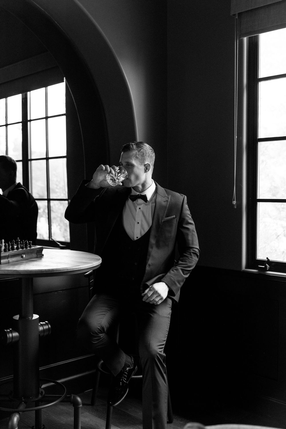 Black and white photo of a well-dressed person in a suit standing by a window in a moody, dimly lit bar setting.