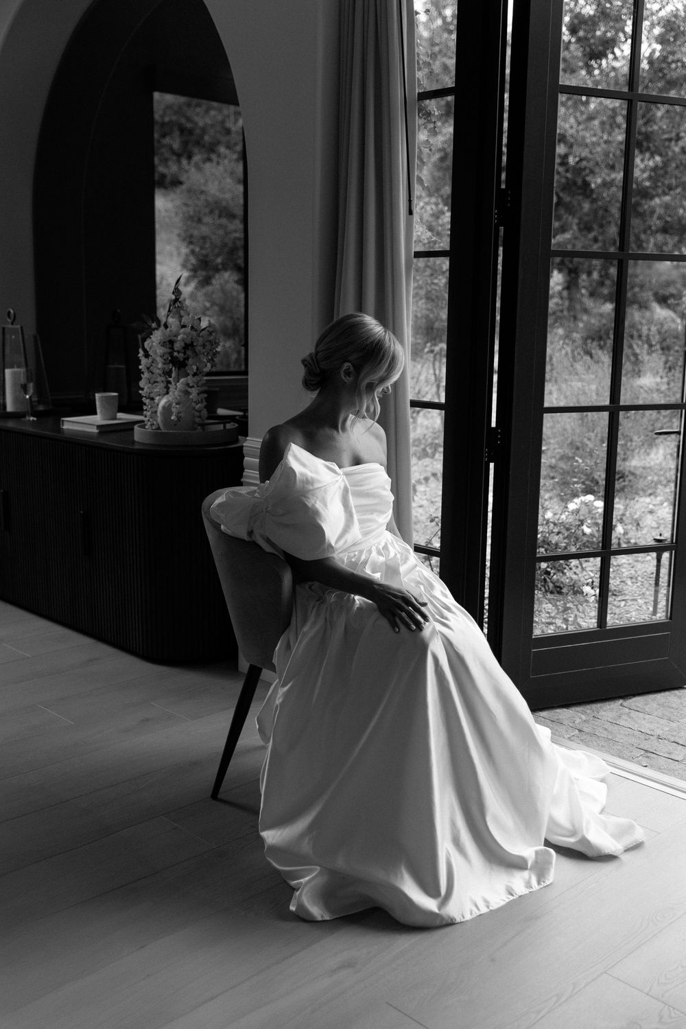A dreamy black and white photo shows a person in a flowing white dress gazing out large windows in contemplative pose.