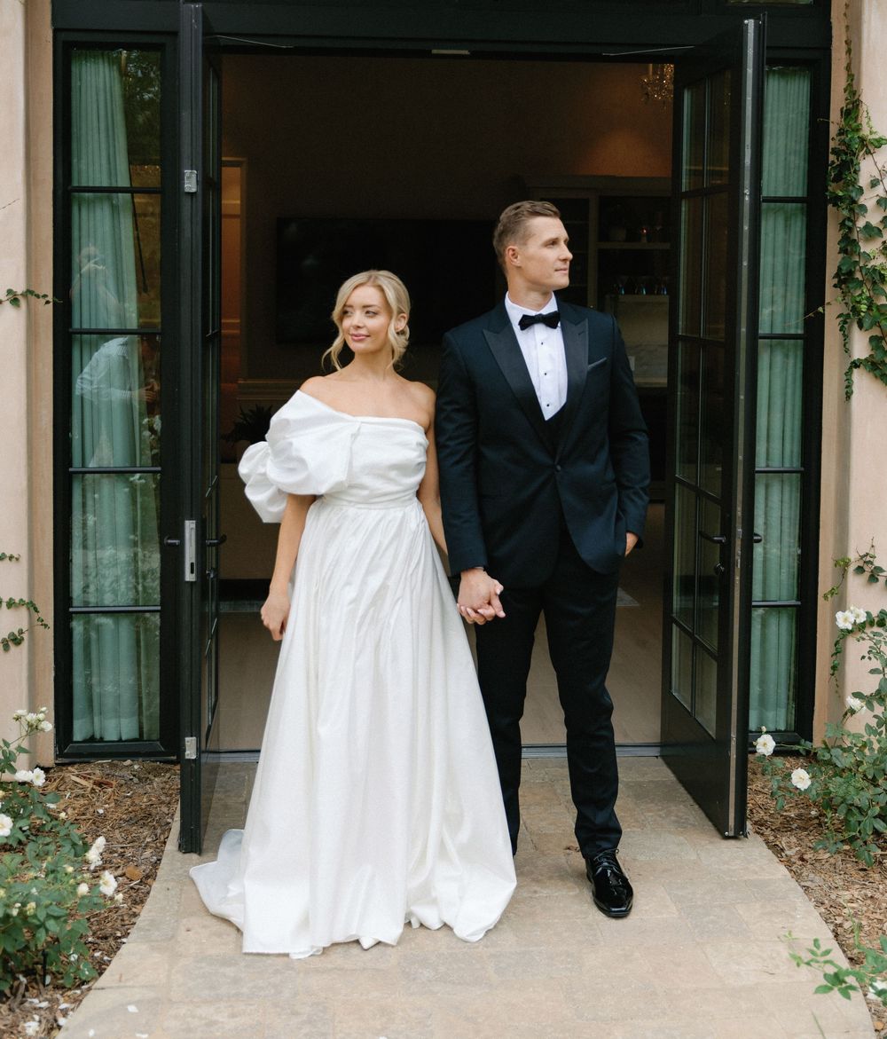 A bride in an off-shoulder white gown stands with a groom in black tuxedo outside elegant French doors.
